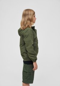Veste bomber verte avec capuche, poignets élastiques et poche zippée, associée à un short cargo vert. Tissu lisse et durable.