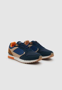 Par de zapatillas deportivas en azul marino, beige y naranja con cordones, tejido de malla y suelas de goma sobre un fondo gris liso.