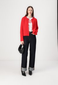 Un cardigan tricoté rouge avec des boutons ronds sur un t-shirt graphique blanc. Un jean foncé avec des ourlets retroussés ; un sac noir et des bottines à talons pointus complètent le look.