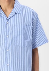 Chemise à manches courtes en bleu clair avec des rayures verticales, dotée d'un devant à boutons et d'une poche poitrine. Fabriquée en tissu léger.