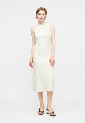 Moteris stovi, vilkinti be rankovių baltą midi ilgio suknelę su gėlių nėrinių detalėmis ir juodus batelius ant aukšto kulno su dirželiais, ant balto fono.