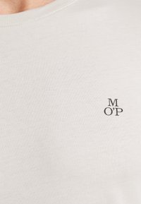 Jasno beżowy T-shirt wykonany z miękkiej tkaniny, z okrągłym dekoltem i małym, ciemnym nadrukiem logo "M OP" po lewej stronie.