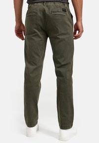 Pantalones verde oliva de corte holgado, con cintura elástica y dos bolsillos traseros. Textura suave con un ligero brillo, combinados con zapatillas blancas.