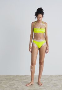 Rumena bikinija z bandeau zgornjim delom s logotipom in ujemajočimi se nizkimi hlačkami. Gladka tekstura, minimalna strojna oprema in čiste oblikovne linije.