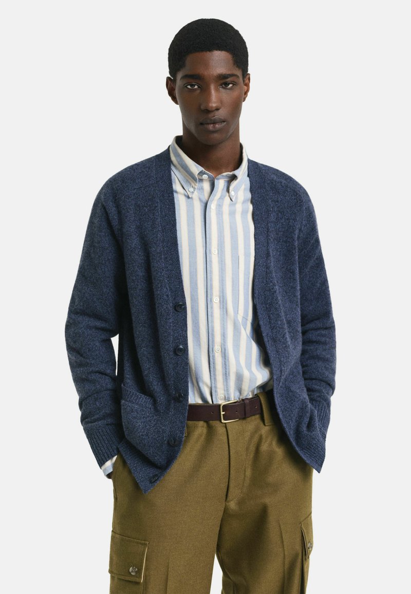 Junger Mann, der einen blauen, geknöpften Cardigan über einem gestreiften Hemd, olivgrünen Cargo-Hosen und einem dunkelbraunen Gürtel trägt, während er mit den Händen in den Taschen steht.