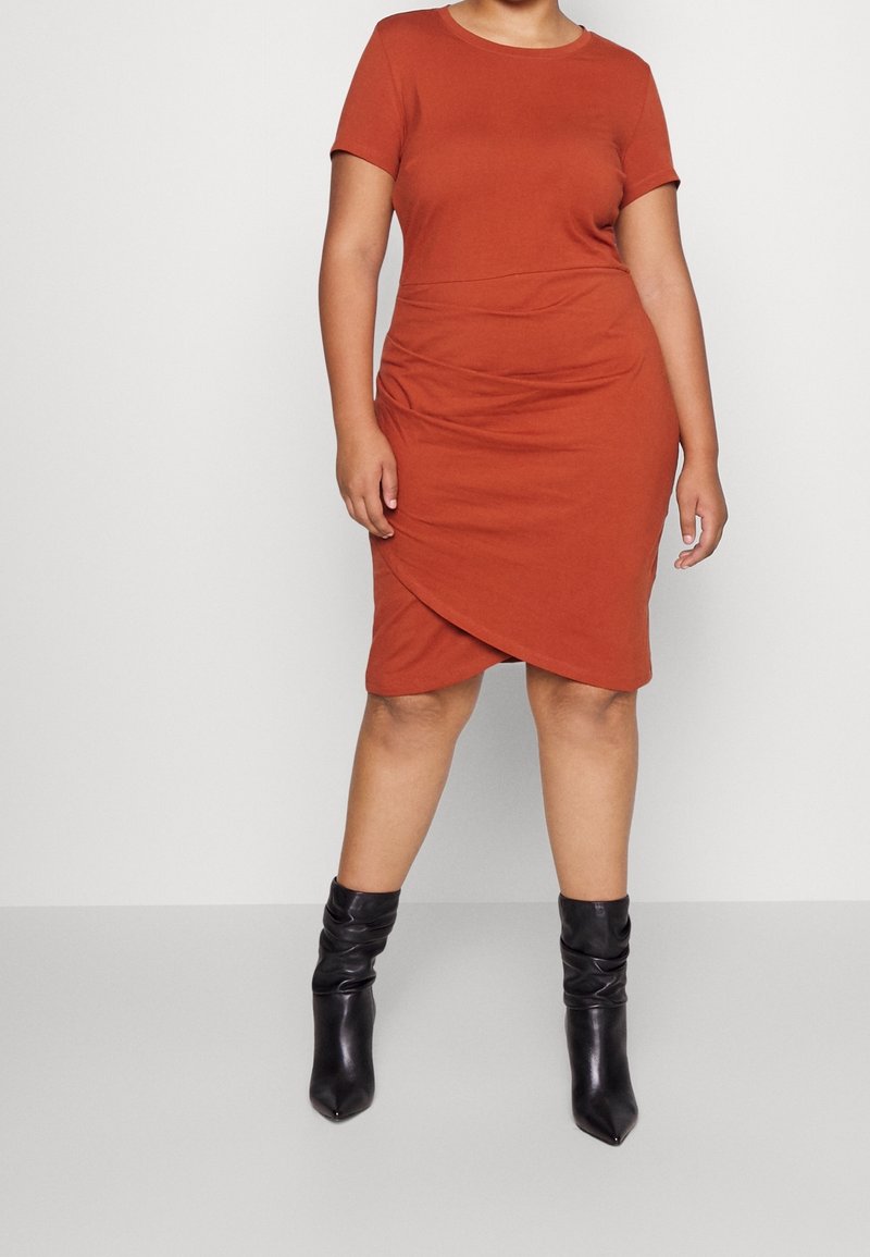 Femme portant une robe portefeuille à manches courtes couleur rouille et des bottes en cuir noires mi-mollet, debout devant un fond uni.