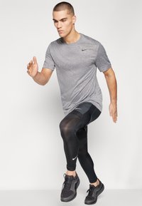 Graues Sportshirt, schwarze Kompressionsleggings mit Nike-Logo und schwarze Laufschuhe. Das Model befindet sich in einer dynamischen Laufpose.