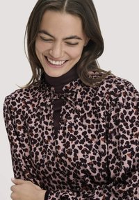 Bluse im Leopardenmuster aus weichem Stoff, mit hohem Kragen und Schleifendetail. Farben sind Pink, Schwarz und einen Hauch von Braun.