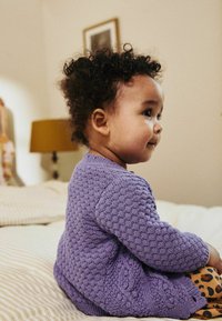 Lila pletena jopica z teksturirano vzorcem, oblečena preko vzorčastih oranžnih in rjavih legic. Otrok sedi na bež preprogi.