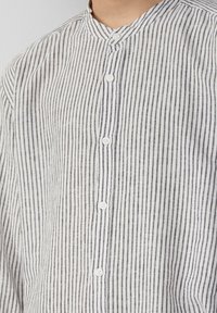 Camicia a righe bianca e grigia. Presenta un colletto a bande, strisce verticali e una patta frontale con bottoni bianchi. Materiale leggero.