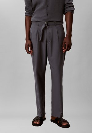 Mann trägt ein graues Langarmshirt, das in graue plissierte Hosen mit einem schmalen Gürtel und schwarzen Sandalen gesteckt ist, vor einem einfarbigen Hintergrund.