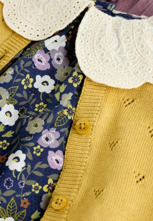 Gelber gestrickter Cardigan mit einem Blumenkleid darunter. Der Cardigan hat einen geschwungenen Kragen und dekorative Knöpfe, die die detaillierten Texturen hervorheben.
