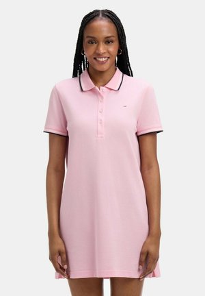 Femme aux cheveux tressés, souriante, portant une robe polo à manches courtes rose clair avec des bordures noires sur le col et les manches.