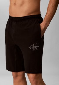 Αρσενικός κορμός και πόδια με μαύρα σορτς Calvin Klein με ελαστική μέση, χέρι στην τσέπη, γκρι φόντο.