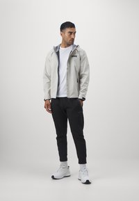 Giacca grigia chiaro con zip e cappuccio, con dettagli neri, abbinata a pantaloni jogger neri, t-shirt bianca e sneaker bianche.