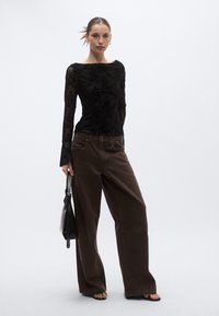 Črno dolga majica s cvetličnimi čipkastimi detajli, v kombinaciji s širokimi rjavimi hlačami. Model drži majhno črno torbico in nosi črne sandale.