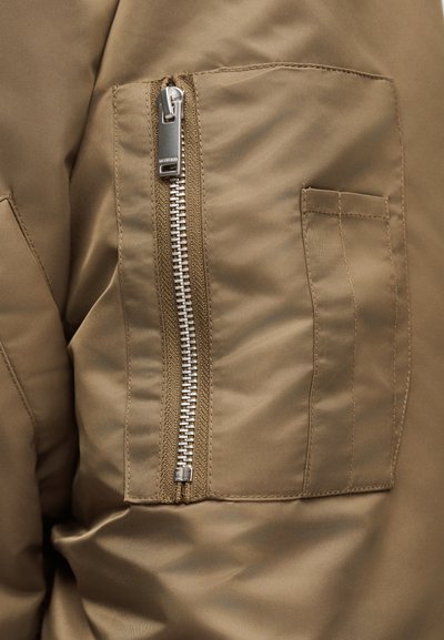 Κοντινό πλάνο μανσετών χακί μπουφάν που δείχνει κάθετο ασημί φερμουάρ δίπλα σε ορθογώνια τσέπη με εμφανή ραφή.