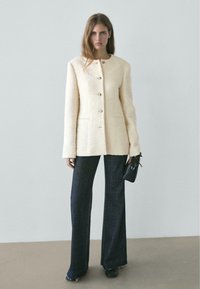 Giacca testurizzata color crema con scollo rotondo, bottoni frontali e due tasche, abbinata a jeans scuri a gamba larga e una piccola borsa nera.