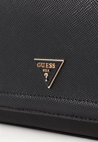 Černý texturovaný syntetický materiál s trojúhelníkovým zlatým logem „GUESS USA“. Obsahuje švy podél okrajů.