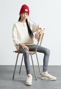 Hvit genser, svart-hvitt rutete leggings, rød strikket lue med logo, og kremfargede sneakers med røde detaljer, sittende på en trestol.