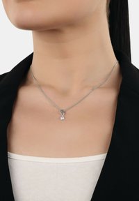 Primo piano di una persona che indossa una sottile collana d'argento con un piccolo pendente circolare che presenta una pietra preziosa rosa, vestita con abiti in bianco e nero.