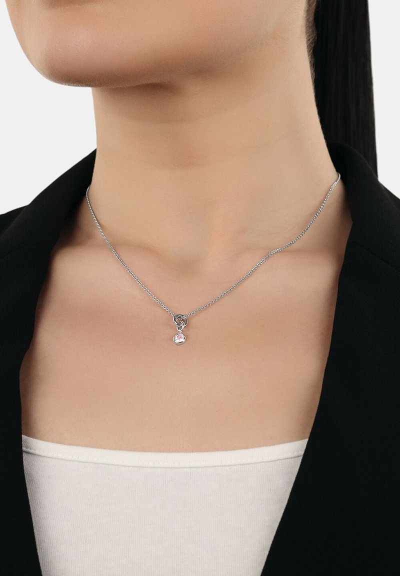Primo piano di una persona che indossa una sottile collana d'argento con un piccolo pendente circolare che presenta una pietra preziosa rosa, vestita con abiti in bianco e nero.