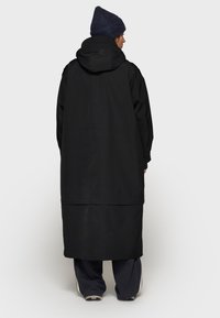 Zwarte lange jas met een capuchon, met een rechte snit en minimalistisch ontwerp. De stof lijkt duurzaam en waterafstotend.