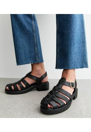 Sandales en cuir noir avec un design à lanières, bout ouvert et fermeture à boucle à la cheville. Talon épais et semelle texturée.
