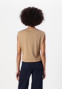 Top sin mangas en tono beige con un corte relajado, cuello redondo y material de algodón suave. La parte trasera presenta un corte a la altura de la cintura y detalles de costura sutiles.