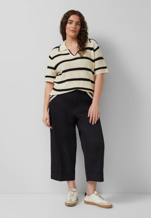 Woman with curly hair wearing a cream and black striped short-sleeve knit top, black wide-leg cropped pants, and white sneakers.