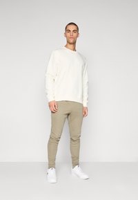 Hanorac crem cu mâneci lungi, asortat cu pantaloni taperi într-o nuanță deschisă de kaki și adidași albi, având un design simplu și fără modele vizibile.