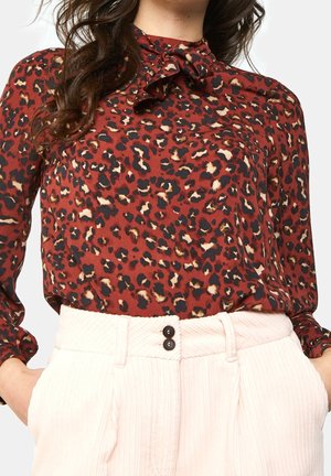 Kobieta w bluzce w cętkowany wzór w kolorze rdzawej czerwieni z zawiązaną kokardą przy szyi oraz w jasnoróżowych spodniach z wysokim stanem z dwoma czarnymi guzikami.