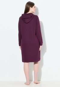 Frau, die barfuß vor einer Wand steht und ein lila Hoodie-Kleid mit langen Ärmeln und knielanger Länge trägt.