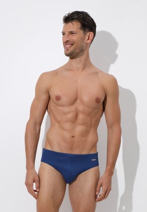 Uomo senza maglietta con capelli corti e scuri, che indossa slip da bagno blu, in piedi e sorridente mentre guarda leggermente a sinistra su uno sfondo bianco uniforme.