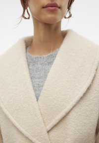 Beige ullkappa med en bred krage, mjuk textur och en grå stickad tröja under. Minimalistisk design utan synlig hårdvara.