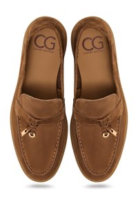Mocasines de ante marrón con punta redonda, detalles de costura decorativa y un pequeño acento de hardware dorado en la parte frontal. Logo de la marca en el interior.