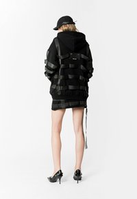 Sudadera oversized negra con acentos de cinta texturizada, combinada con una mini falda negra a juego. Los accesorios incluyen una gorra negra y tacones brillantes negros.