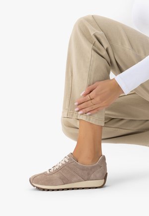 Beżowy zamszowy but sportowy z białymi sznurówkami na nodze, zestawiony z beżowymi spodniami i białą bluzką z długim rękawem, ręka spoczywająca na kostce.