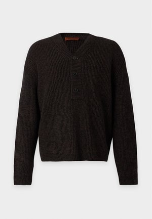 Man draagt een donkere geribde trui met knopen, zwarte broek en zwarte schoenen, staand tegen een effen witte achtergrond.