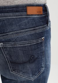 Jeans de mezclilla azul con un parche de cuero con la marca "edc", costuras en contraste y un sutil detalle bordado en el bolsillo trasero.