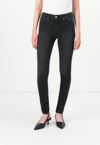 Egy nő fekete skinny farmert és fekete, hegyes orrú szandált visel, egy sima fehér háttér előtt áll.