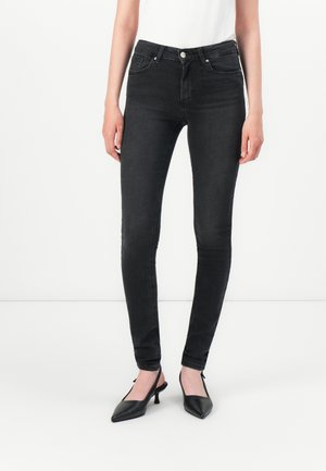 Woman wearing black skinny jeans and black pointed-toe slingback heels, standing against a plain white background.