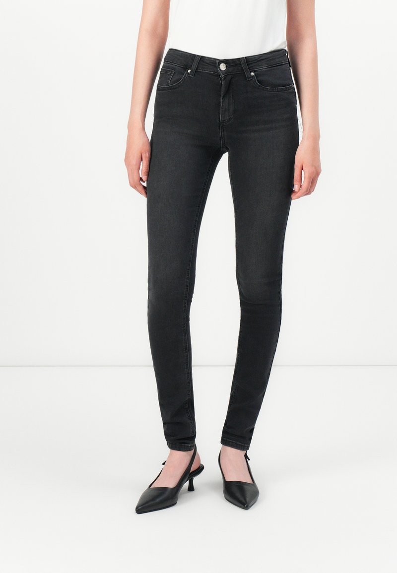 Egy nő fekete skinny farmert és fekete, hegyes orrú szandált visel, egy sima fehér háttér előtt áll.