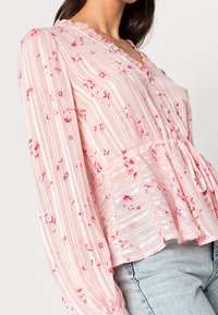 Blusa floreale rosa con strisce verticali, colletto a volant e maniche lunghe. Tessuto leggero con vita a peplum e bottoni decorativi.