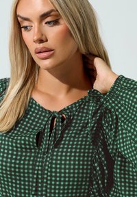 Vihreä ja musta ruudullinen pusero, jossa on pyöreä kaula-aukko, nauhakiinnitys ja kokoontuneet hihat. Sileä, kevyt kangas, jolla on tekstuurinen ilme.