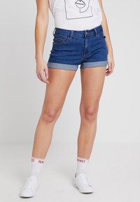 Shorts de mezclilla con un lavado azul oscuro, dobladillos enrollados, bolsillos frontales y un diseño ajustado, combinados con una camiseta gráfica blanca y zapatillas blancas.