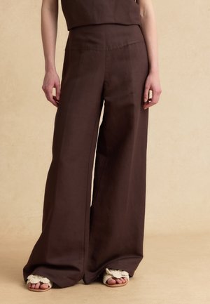 Mujer que lleva pantalones anchos y sueltos de color marrón oscuro y sandalias tejidas color crema con los dedos abiertos, de pie frente a un fondo neutro.