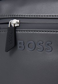 Melnā ādas maciņš ar iegravētu "BOSS" logo, gluda tekstūra, sudraba metāla rāvējslēdzis un gumijas rāvējslēdzēja vilcējs ar rakstainu virsmu.