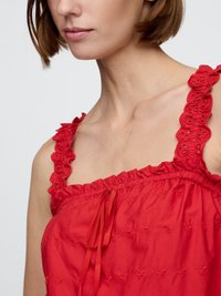 Prim-plan al unei femei care poartă un top roșu fără mâneci, cu bretele din dantelă cu volane și un decolteu gathered cu o panglică legată.