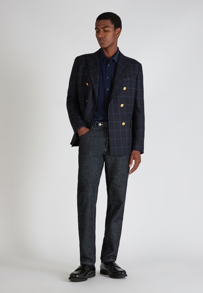 Mann in karierter, dunkler Jacke mit goldenen Knöpfen, navyblauem Hemd, dunklen Jeans und schwarzen Schuhen, steht mit einer Hand in der Tasche.